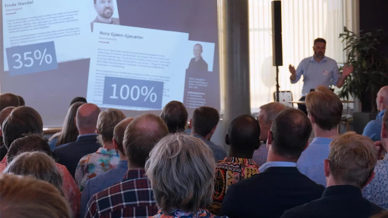 Publikum følger med på et foredrag i et fullsatt lokale, med en foredragsholder ved en skjerm